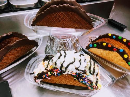 Four ice cream tacos in waffle cones, topped with chocolate, sprinkles, and colorful candies, displayed on a silver tray.