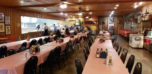 Interior of a rustic restaurant with checkered tablecloths, empty tables, and a visible kitchen area in the background.