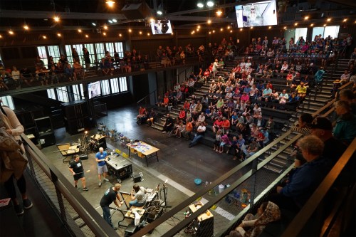 A large audience watches a live demonstration on stage in a modern auditorium with multiple screens and bright lighting.
