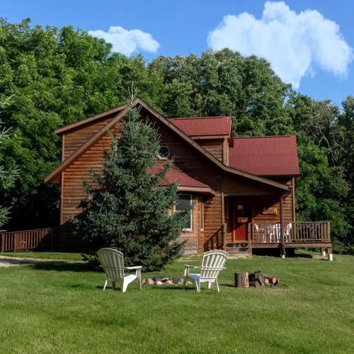 A wooden cabin with a red roof surrounded by trees, featuring a porch and two white chairs in the foreground.