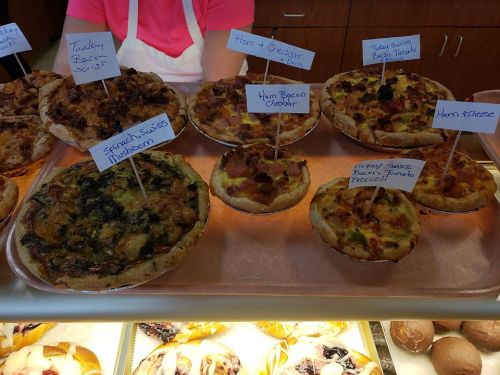 A display of various savory pastries, including ham and cheese, spinach mushroom, and turkey bacon options, labeled on signs.