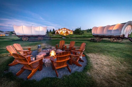 A cozy campfire surrounded by wooden chairs, with vintage wagons and a cabin in the background at dusk.