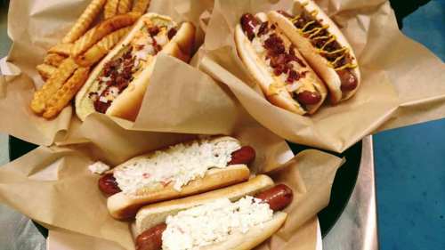 A tray of hot dogs with various toppings and a side of crinkle-cut fries, all served in paper wrappers.