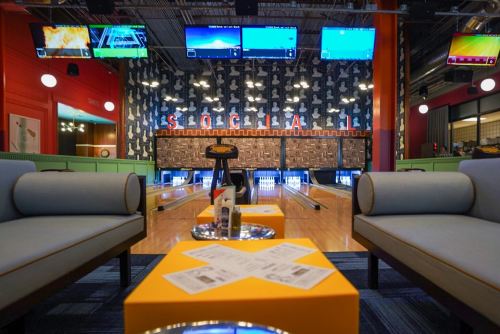 A vibrant bowling alley lounge with blue sofas, yellow tables, and multiple screens displaying bowling games.