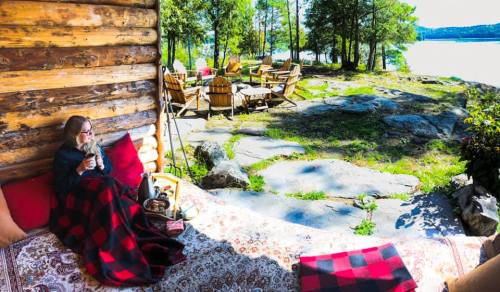 A woman relaxes on a cozy outdoor seating area by a lake, surrounded by trees and rustic furniture.