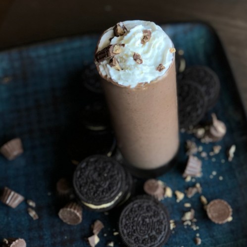 A tall chocolate milkshake topped with whipped cream and chocolate pieces, surrounded by Oreo cookies and candy.