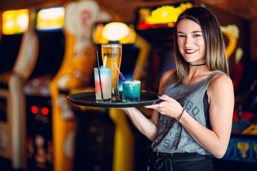 A smiling woman holds a tray of colorful cocktails in a retro arcade setting.