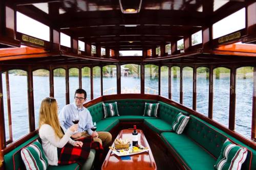 A couple enjoys wine and snacks on a cozy boat with green seating, surrounded by water and trees.