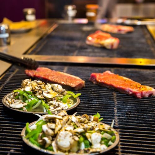 Grilled steaks and sautéed vegetables on a barbecue grill, with smoke rising in the background.