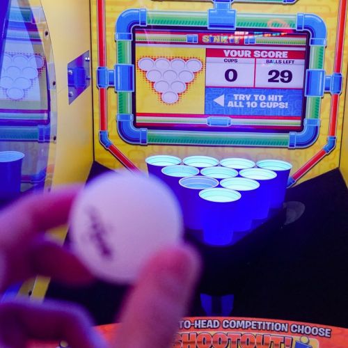A hand holds a ping pong ball in front of a colorful arcade game displaying a score of 0 and 29 balls left.