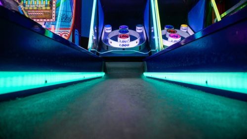 A close-up view of an arcade game lane with glowing blue lights and scoring buttons in the background.