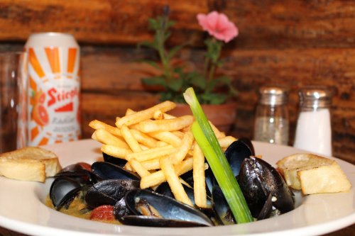 A plate of mussels topped with fries and green onion, accompanied by bread, a soda can, and a small plant in the background.