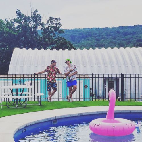 Two friends in colorful shirts stand by a pool with a pink flamingo float, surrounded by greenery and a white building.