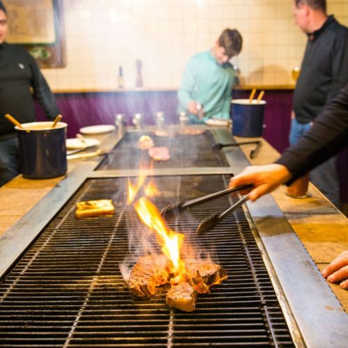 A grill with steaks cooking over flames, while people prepare food in a cozy kitchen setting.