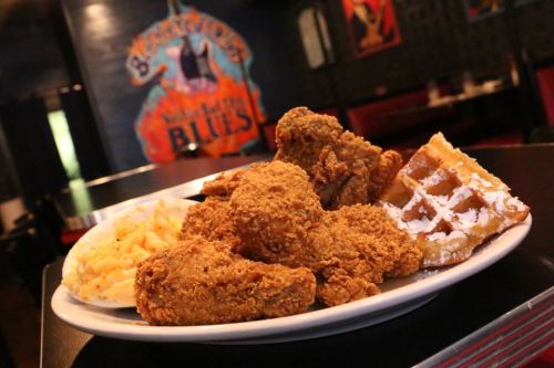 A plate of crispy fried chicken, a waffle, and a side of macaroni and cheese, set against a colorful restaurant backdrop.