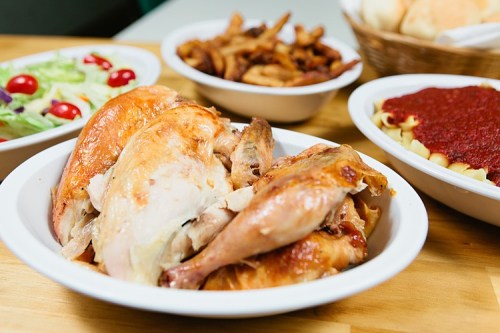 A platter of roasted chicken with sides of salad, pasta with sauce, and crispy fried onions on a wooden table.