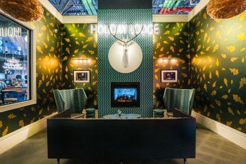 Cozy interior with patterned wallpaper, a fireplace, two chairs, and a mounted deer head, illuminated by neon signage.