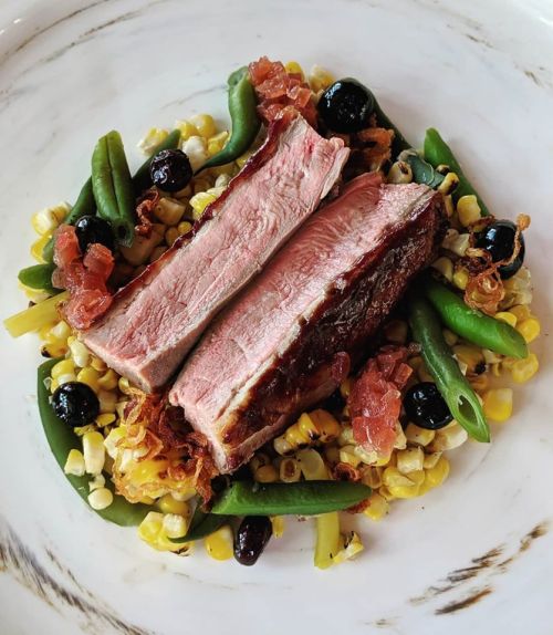 A plate of sliced steak on a bed of corn, green beans, and garnished with crispy onions and black olives.