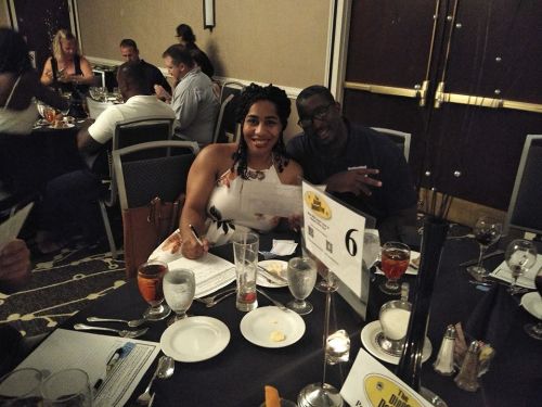 A couple smiles at a dining table with drinks and papers, surrounded by other guests in a restaurant setting.