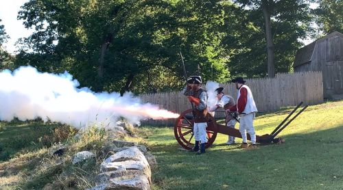 Historical reenactors fire a cannon, creating a cloud of smoke, with a wooden fence and trees in the background.
