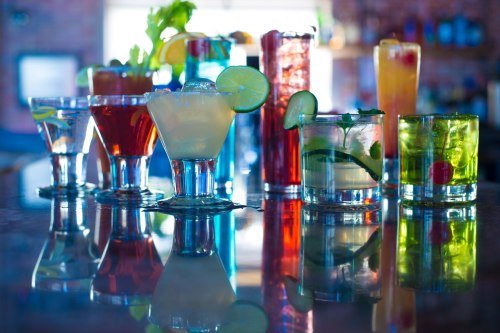 A colorful array of cocktails displayed on a bar, reflecting in the surface, with garnishes like lime and celery.