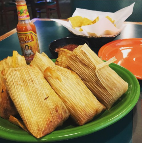 A plate of tamales with a bottle of hot sauce, a bowl of salsa, and a basket of tortilla chips in the background.