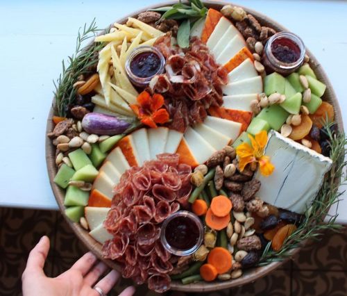 A colorful charcuterie board with cheeses, meats, fruits, nuts, and dips, garnished with herbs and edible flowers.