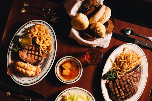 Two plates of steak with fries, a bowl of soup, and a basket of assorted bread rolls on a wooden table.