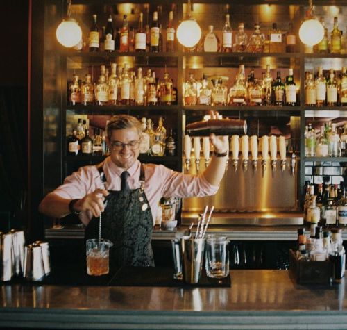A bartender in a pink shirt and apron mixes a drink at a well-stocked bar with various bottles in the background.