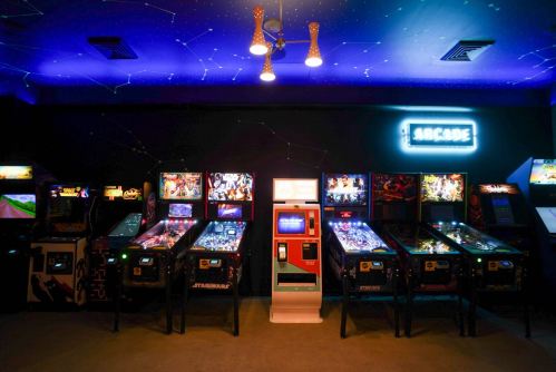 A vibrant arcade with various pinball machines and a neon "Arcade" sign against a starry ceiling.
