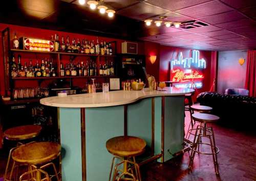 A stylish bar with a blue counter, wooden stools, shelves of liquor, and a neon sign in a cozy, dimly lit space.