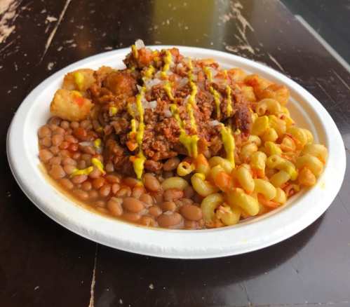 A plate of food featuring macaroni, baked beans, tater tots, and a meat topping drizzled with mustard.
