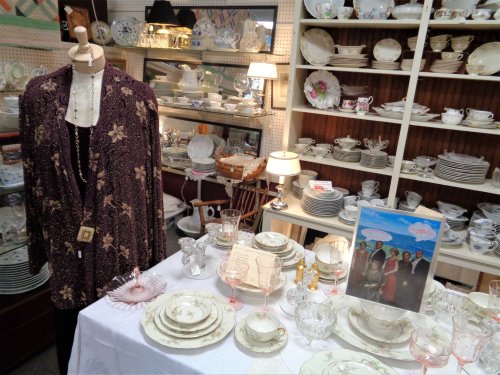 A vintage display featuring a mannequin in a patterned shawl, elegant tableware, and decorative glassware.