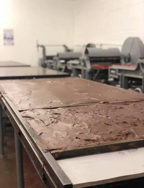 A large slab of melted chocolate on a metal table in a factory setting, with machinery in the background.