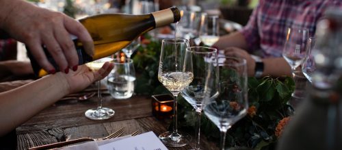 A hand pours white wine into a glass at a beautifully set dining table with greenery and candles.