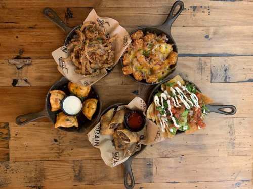 Four cast iron dishes filled with various appetizers, including nachos, fried dough, and loaded tater tots on a wooden table.