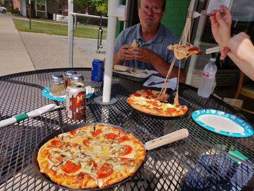 A table with two pizzas, drinks, and people enjoying a meal outdoors on a sunny day. Cheese stretches from a slice.