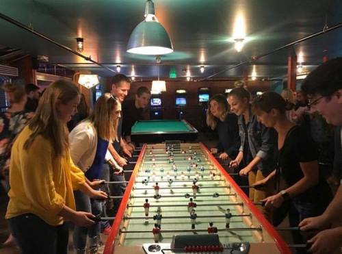 A group of people playing foosball in a lively arcade setting, with bright lights and arcade games in the background.