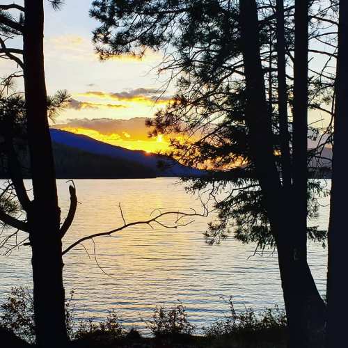 Sunset over a calm lake, framed by silhouetted trees and mountains in the background.