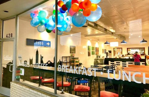 A café window decorated with colorful balloons, showcasing a cozy interior with tables and a coffee counter.