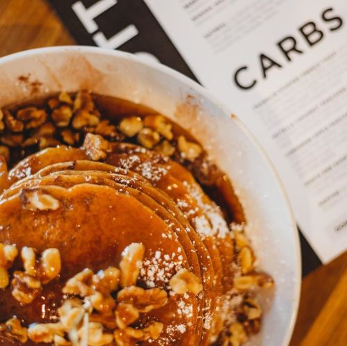 A bowl of stacked pancakes topped with walnuts and syrup, next to a menu featuring the word "CARBS."