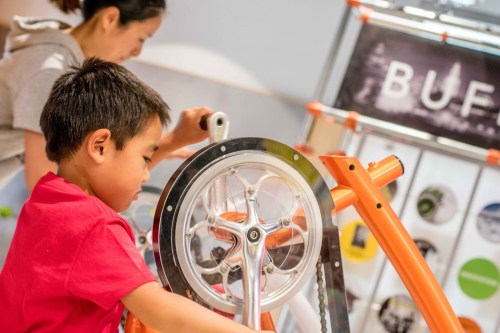 A child in a red shirt interacts with a colorful bike-like exhibit while an adult observes in the background.