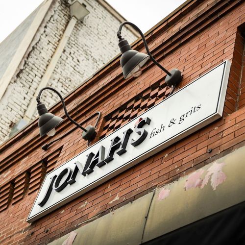 Sign for "Jonah's Fish & Grits" on a brick building, featuring decorative lighting above.