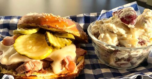 A savory sandwich with ham, melted cheese, and pickles, alongside a bowl of creamy fruit salad on a checkered cloth.
