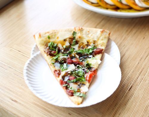 A slice of pizza topped with herbs, tomatoes, and cheese on a paper plate, set on a wooden table.