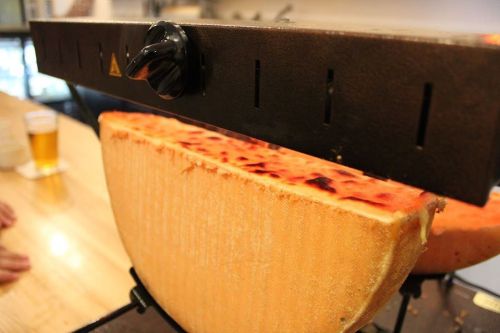 A wheel of cheese melting under a grill, with a glass of beer in the background on a wooden table.