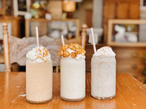 Three milkshakes in tall glasses, topped with whipped cream and various toppings, on a wooden table.