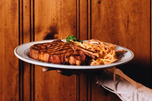 A hand holds a plate with a grilled steak and crispy onion strings, set against a wooden background.
