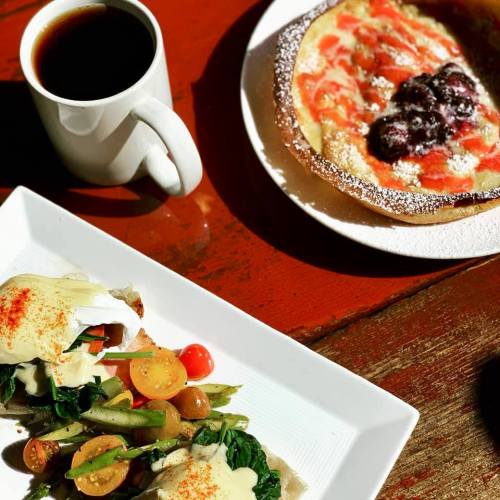 A plate of eggs Benedict with vegetables, alongside a fruit tart and a cup of coffee on a wooden table.