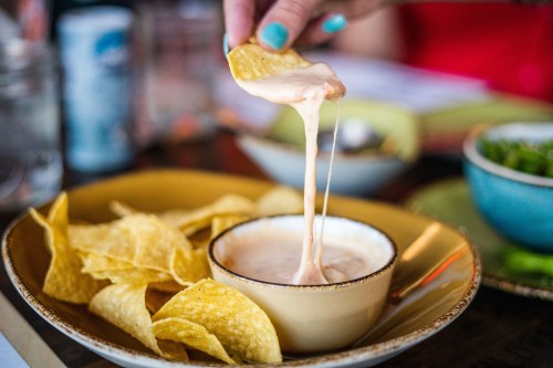 A hand dips a tortilla chip into a bowl of creamy cheese sauce, surrounded by crispy chips on a plate.